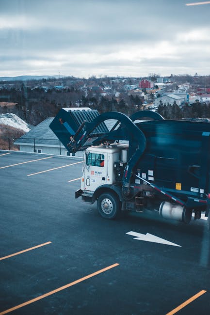 A white waste collection truck with hydraulic lifting arms is parked on an empty, well-marked rooftop parking area, which has yellow lane markings. The vehicle is positioned to the right side of the i