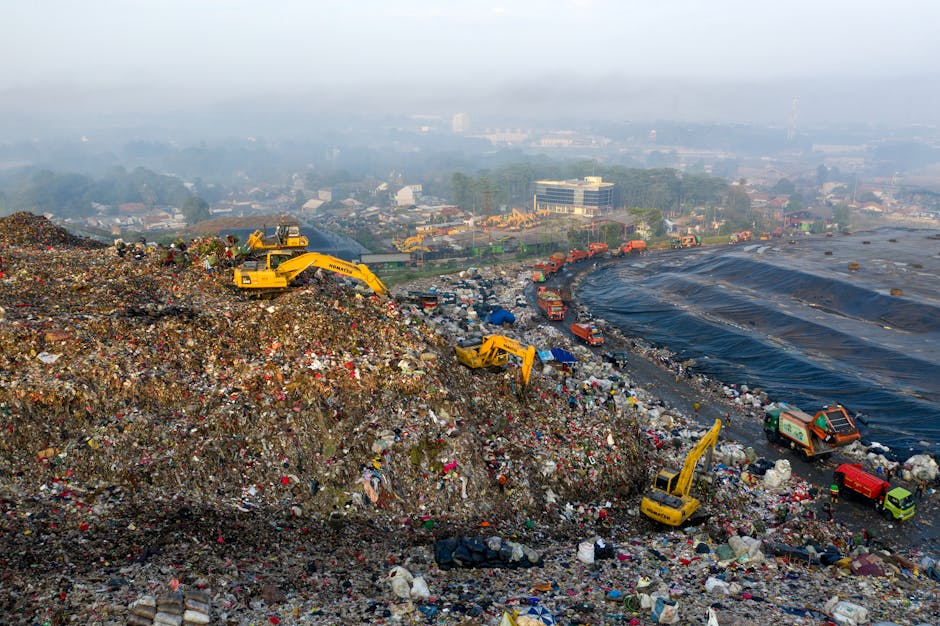 A large landfill site with a prominent mound of mixed waste materials, including plastic bottles, bags, and various refuse, scattered unevenly across the terrain. Several yellow excavators are activel